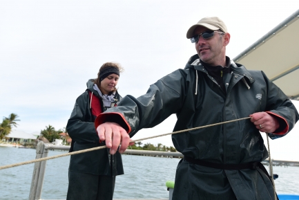 : A man in a black jacket with sunglasses and a young woman stand on a boat. The man holds a line of rope in front of him.