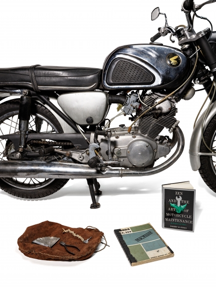 Black motorcycle with two books and a leather bag on ground next to it.