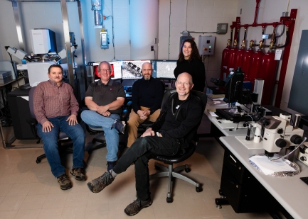 A group of four people sit in office chairs in a scientific lab.