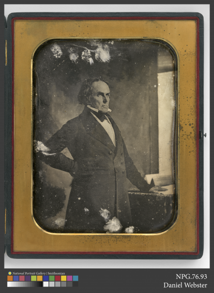 Antique black and white image of a man from the waist up wearing a suit, standing in front of a window.