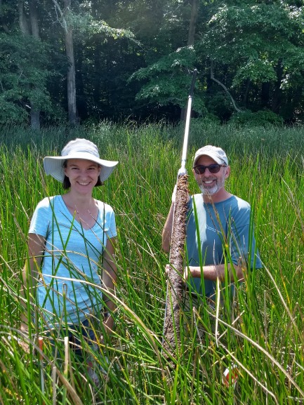A man and a woman wearing short sleeve shirts and outdoors attire stand in field of tall green gas holding large stick with soil