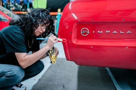 A young person wearing jeans and short sleeve shirt crouches next to end of red Impala.
