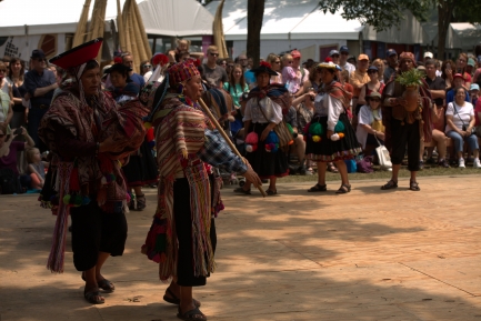 A man in a vibrant colorful outfit plays a long wooden flute in front of a crowd.
