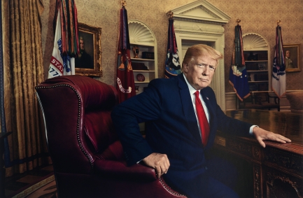 Man in black suit and red tie sits at a dark wooden desk in the Oval Office.