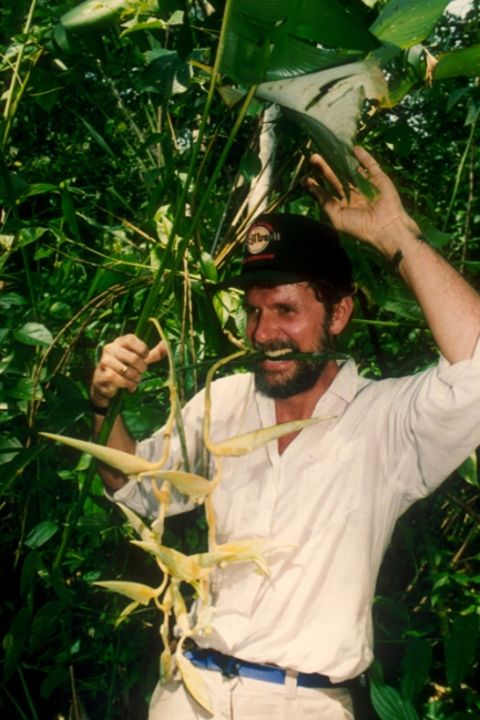 Man in button down and a black cap stands in the forest holding a leaf sample.