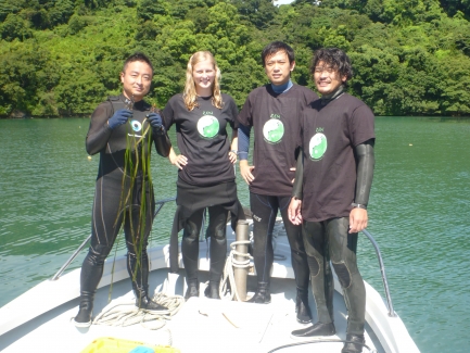 Four people in wet suits pose on edge of a boat on water