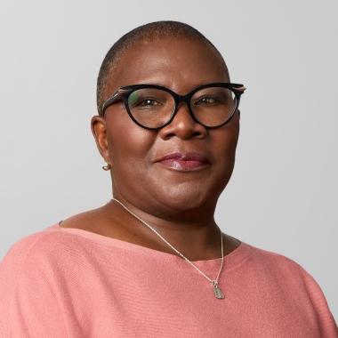 Woman with hair cropped close to head, wearing cat eye glasses and a pink top, smiles toward camera.