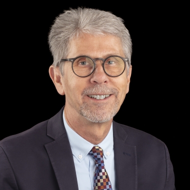 Photo of a man, from chest up, with short white hair and round glasses in a suit and tie.