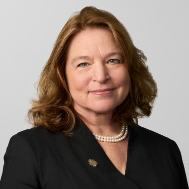 Woman with short blond hair, wearing a black suit and pearls, smiles toward camera.