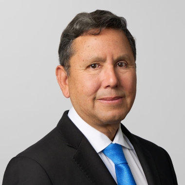Man with short black hair, a black blazer and bright blue tie smiles toward camera.